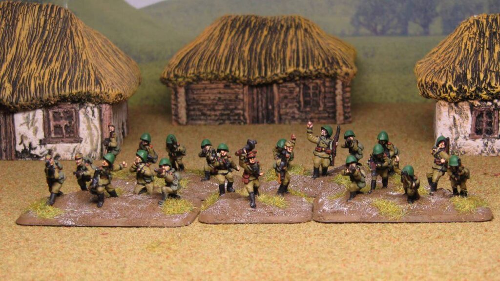 Historical wargaming infantry units on custom textured bases with mud and static grass effects near rural buildings