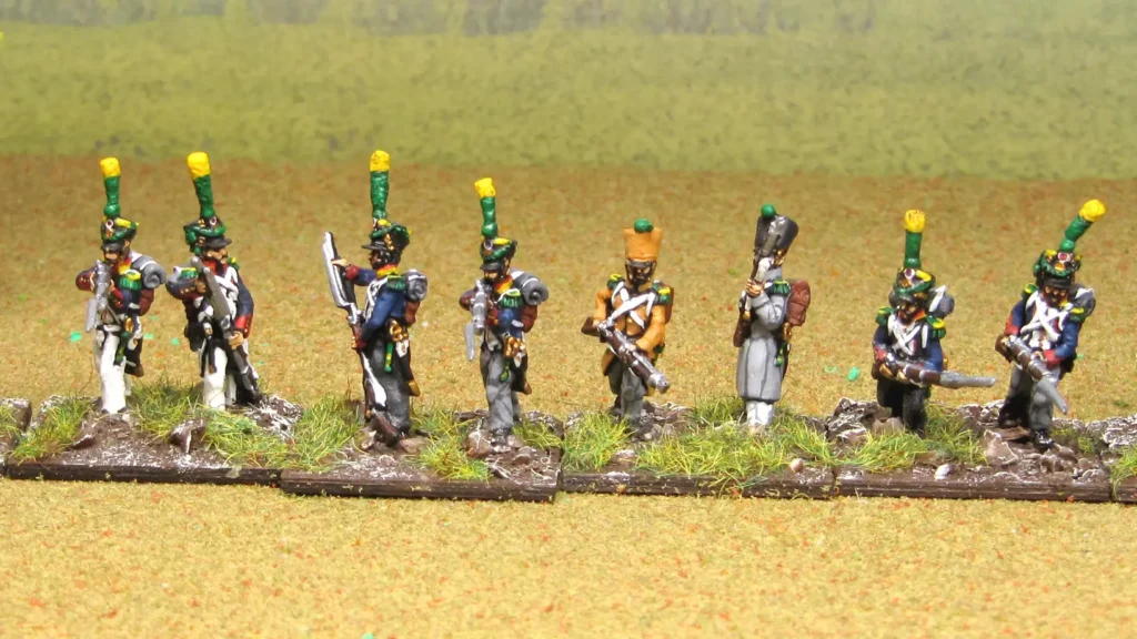 A rank of painted 28mm Napoleonic infantry miniatures standing on matching textured bases with earth, small rocks, and static grass tufts