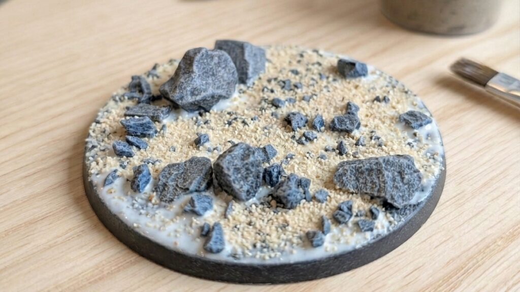 Close-up of an unfinished miniature base with applied PVA glue, fine sand, and small stones before priming and painting